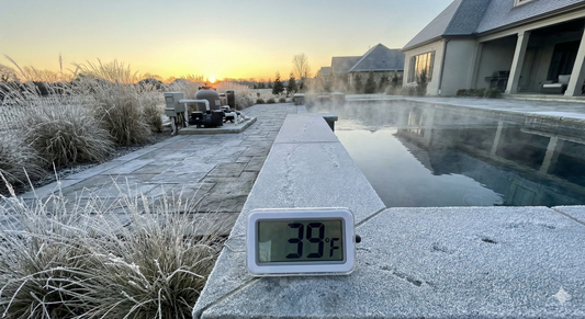 Luxury swimming pool deck covered in morning frost with steam rising from the water. A digital thermometer in the foreground reads 39 degrees Fahrenheit, illustrating the risk of swimming pool freeze damage before temperatures hit freezing.